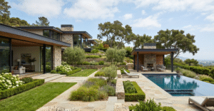 A wide-angle view of a sophisticated, modern residential landscape design, featuring layered stone terraced gardens with hydrangeas and lavender, a geometric lawn path, an infinity pool with chaise lounges, and a rustic stone fireplace pavilion, all set against rolling green hills under a bright sky.